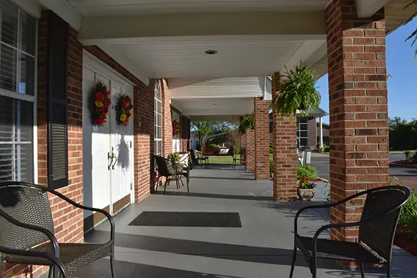Front porch of Caremoor Assisted Living with wicker chairs, hanging plants, and seasonal wreaths on brick columns