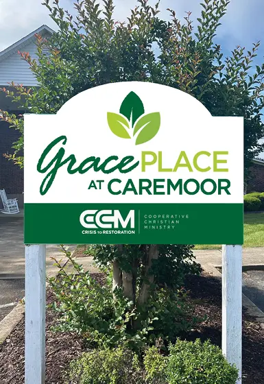 Grace Place at Caremoor sign displaying the logo of CCM, with greenery and trees in the background, representing a place of care and support.